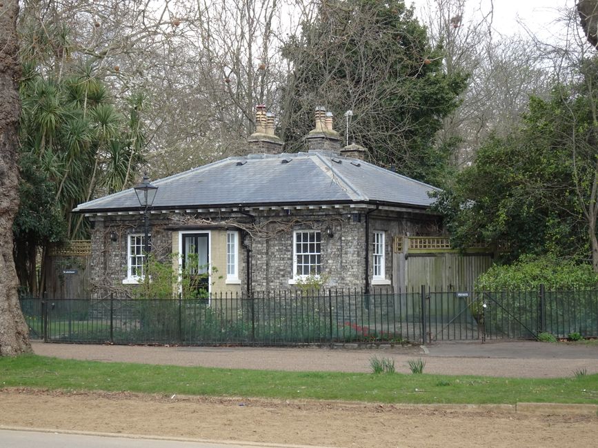 A small, circular brick house with a grey tiled, octagonal roof featuring multiple chimneys, situated behind a black metal fence. The house has white-framed windows and a white door with glass panels, partially covered by climbing plants. The garden in front of the house contains green grass and bushes, with a paved path leading to the entrance. Surrounding the property are tall trees and dense foliage, indicating a suburban or rural setting. The scene appears to be captured during daylight hours with overcast weather, and the house is part of a property that may be involved in home relocation or furniture transport activities, possibly related to the services of Man With a Van Friern Barnet.