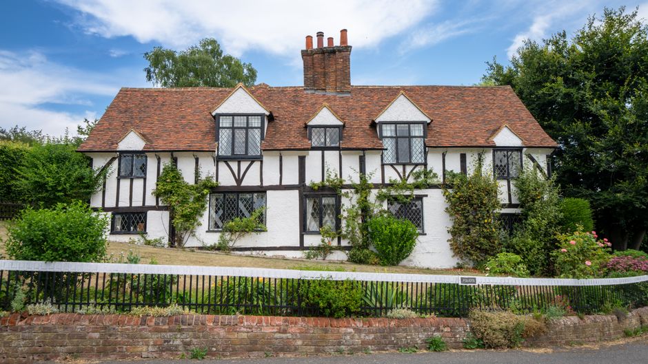 A two-storey, historic Tudor-style cottage with white walls and dark wooden framing, situated in a green garden with various shrubs and climbing plants along the exterior walls. The roof is covered with reddish-brown tiles, and there are four prominent chimney stacks extending above the roofline. The house features multiple black-framed casement windows with diamond-shaped leaded glass panes, positioned symmetrically across the facade. The garden is enclosed by a low brick wall and a black metal fence, with well-maintained flowering plants and bushes along the perimeter. The scene is set outdoors during daytime with a partly cloudy sky, and the environment suggests the initial stages of a home relocation or furniture transport process, as referenced by a moving services provider such as Man With a Van Friern Barnet, supporting local removals and house moving logistics.