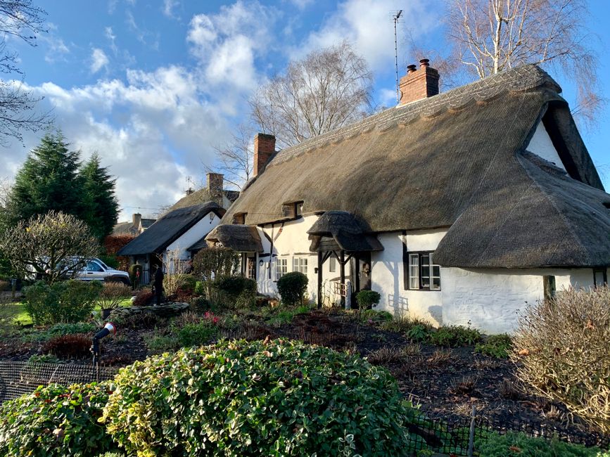A small, circular brick house with a grey tiled, octagonal roof featuring multiple chimneys, situated behind a black metal fence. The house has white-framed windows and a white door with glass panels, partially covered by climbing plants. The garden in front of the house contains green grass and bushes, with a paved path leading to the entrance. Surrounding the property are tall trees and dense foliage, indicating a suburban or rural setting. The scene appears to be captured during daylight hours with overcast weather, and the house is part of a property that may be involved in home relocation or furniture transport activities, possibly related to the services of Man With a Van Friern Barnet.