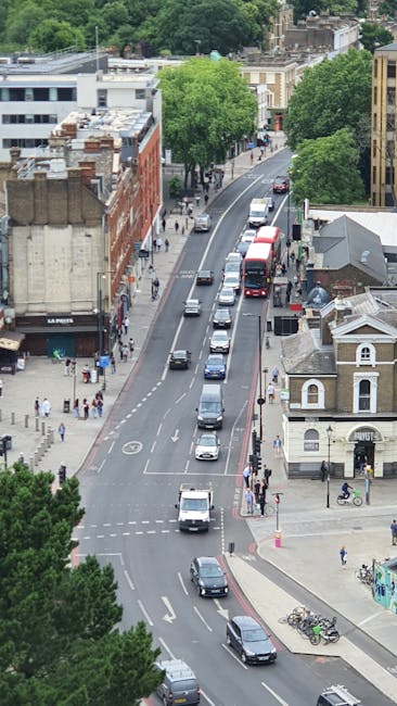An aerial view of Friern Barnet Road showing a busy street scene with multiple vehicles, including cars, vans, and a bus, traveling along a curved road lined with trees and a mixture of modern and traditional buildings. Pavements on either side of the road are occupied by pedestrians, some walking and others waiting at crossings. Bicycles are parked near a small building at street corner, with a few cyclists riding along the pavement. The scene features lush green foliage from the trees, and daylight illuminates the street, creating a vibrant atmosphere suitable for discussions about home relocation or furniture transport during house removals, as typically coordinated by companies like Man With a Van Friern Barnet.