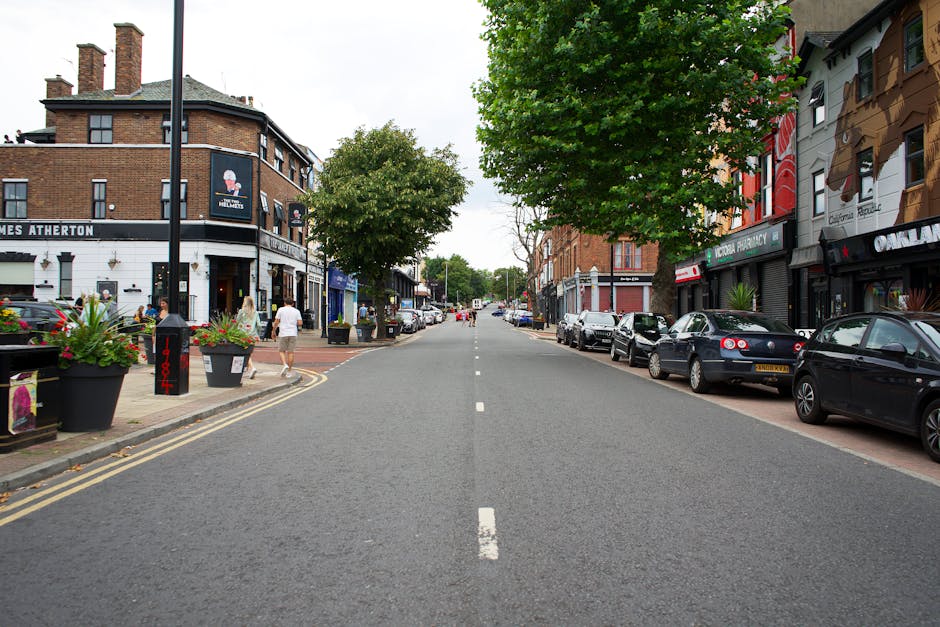An aerial view of Friern Barnet Road showing a busy street scene with multiple vehicles, including cars, vans, and a bus, traveling along a curved road lined with trees and a mixture of modern and traditional buildings. Pavements on either side of the road are occupied by pedestrians, some walking and others waiting at crossings. Bicycles are parked near a small building at street corner, with a few cyclists riding along the pavement. The scene features lush green foliage from the trees, and daylight illuminates the street, creating a vibrant atmosphere suitable for discussions about home relocation or furniture transport during house removals, as typically coordinated by companies like Man With a Van Friern Barnet.
