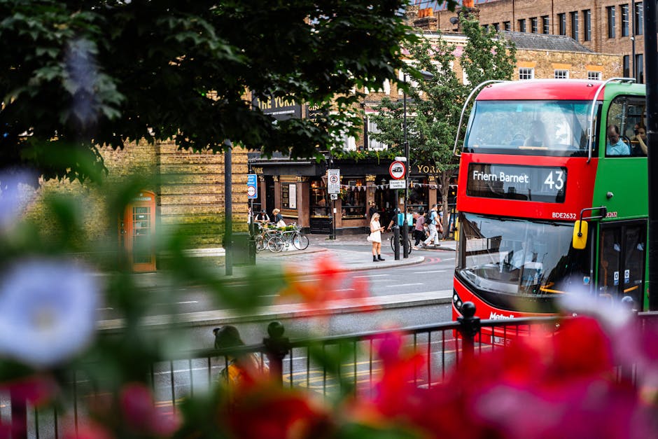 A view from a residential property window showing a busy urban street scene near Arnos Grove station in Friern Barnet. In the foreground, out-of-focus colorful flowers and green leaves partially obscure the scene. Beyond the foliage, a red and green double-decker bus with the route number 43 is seen parked or moving along the street, with the digital display indicating 'Friern Barnet'. The street is lined with pedestrians crossing or standing at the corner, some carrying shopping bags or chatting, and bicycles are parked near a lamppost. Behind the bus, a cluster of buildings with varied brick facades, some with signage, host retail shops and cafes, contributing to the lively atmosphere typical of city centre surroundings. The overall scene captures transportation, foot traffic, and urban life, reflecting the typical context in which a house removal or moving service from Man With a Van Friern Barnet might operate during a home relocation in this area.