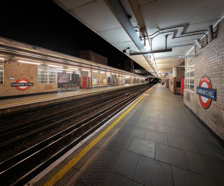 Inside the Harrow & Hillingdon underground station platform, showing a long, empty platform with tracks in the center, illuminated by overhead lighting. The platform is lined with light-colored tiled flooring and features station signage on brick walls, including the Harrow & Hill roundel logos. A row of large windows and benches are visible along one side of the platform, with a designated waiting room sign overhead. The ceiling has metal pipes and electrical conduits, with some visible ventilation ducts. The scene appears prepared for a home relocation or furniture transport operation, with no visible moving equipment or boxes in this particular shot, focusing instead on the station environment for potential loading or unloading of items during a house removals process. Man With a Van Friern Barnet occasionally conducts such loading and packing activities at locations like this station during furniture transport and moving logistics.